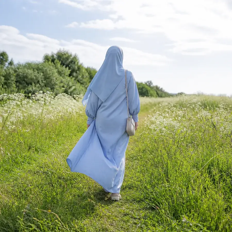 A woman in a flowing pale blue abaya and hijab walking through a sunlit wildflower meadow towards open space